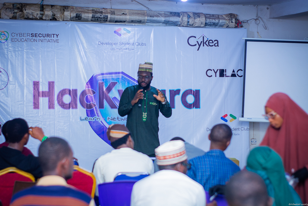Aliyu speaking at a cybersecurity conference, presenting to an engaged audience with Cybersecurity Education Initiative and CyBlac banners visible in the background