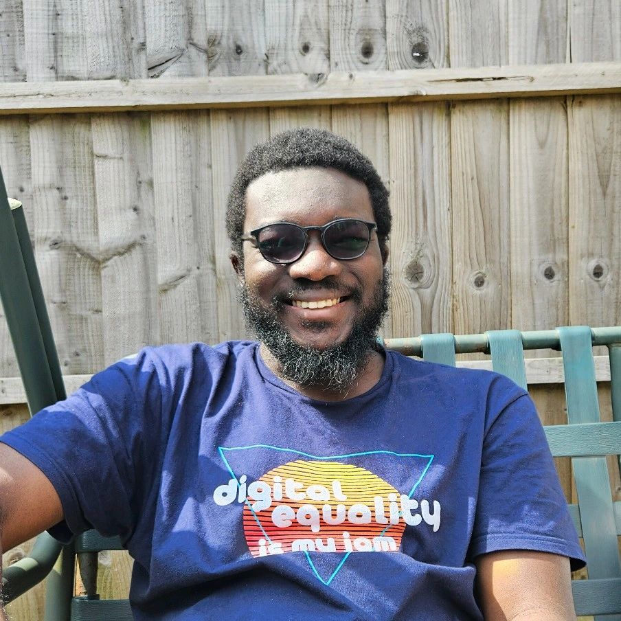 Portrait of Aliyu wearing glasses and a blue t-shirt with 'digital equality' text, smiling in an outdoor setting with a wooden fence background